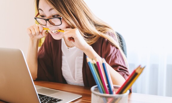 Przy biurku siedzi młoda dziewczyna w okularach. Zagryza ołowek. Przed nią stoi laptop obok pojemnik z kolorowymi kredkami